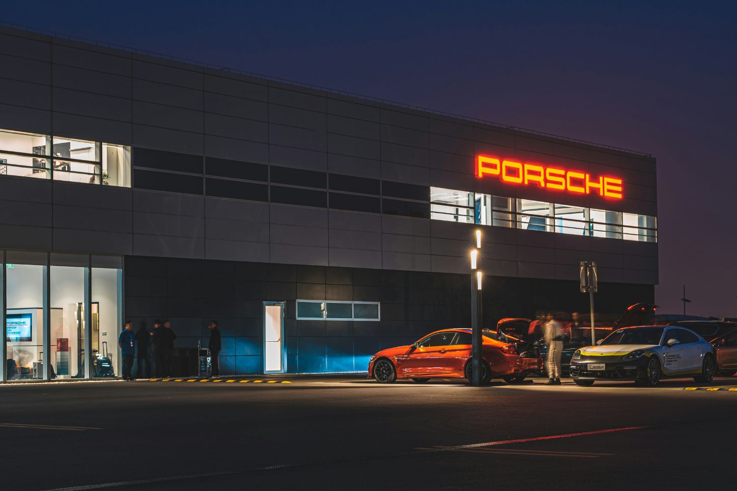 Several Porsche cars parked outside a modern Porsche dealership building with illuminated sign lit up at night